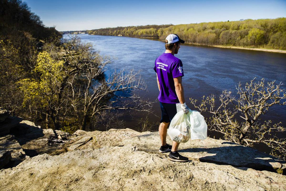A student stands along the shore of the Mississippi River with gloves and a bag of trash.