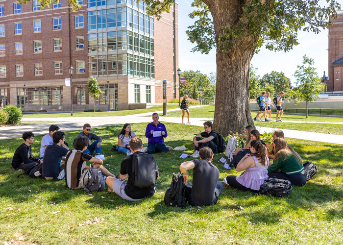 students and professor hold class outside in the shade of a tree
