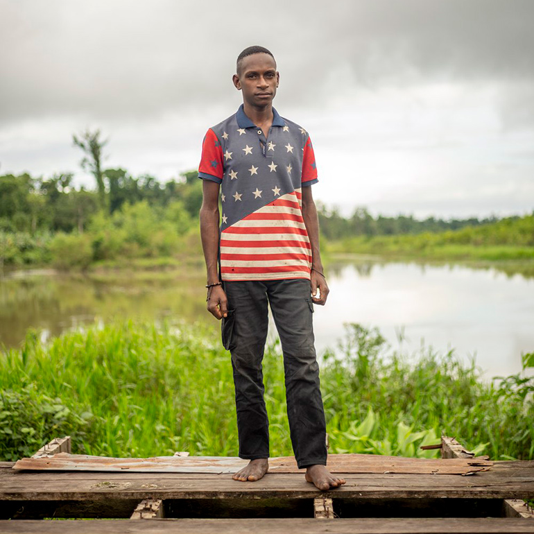 An Indonesian man stands on a wooden dock in an American Flag shirt with water in the background