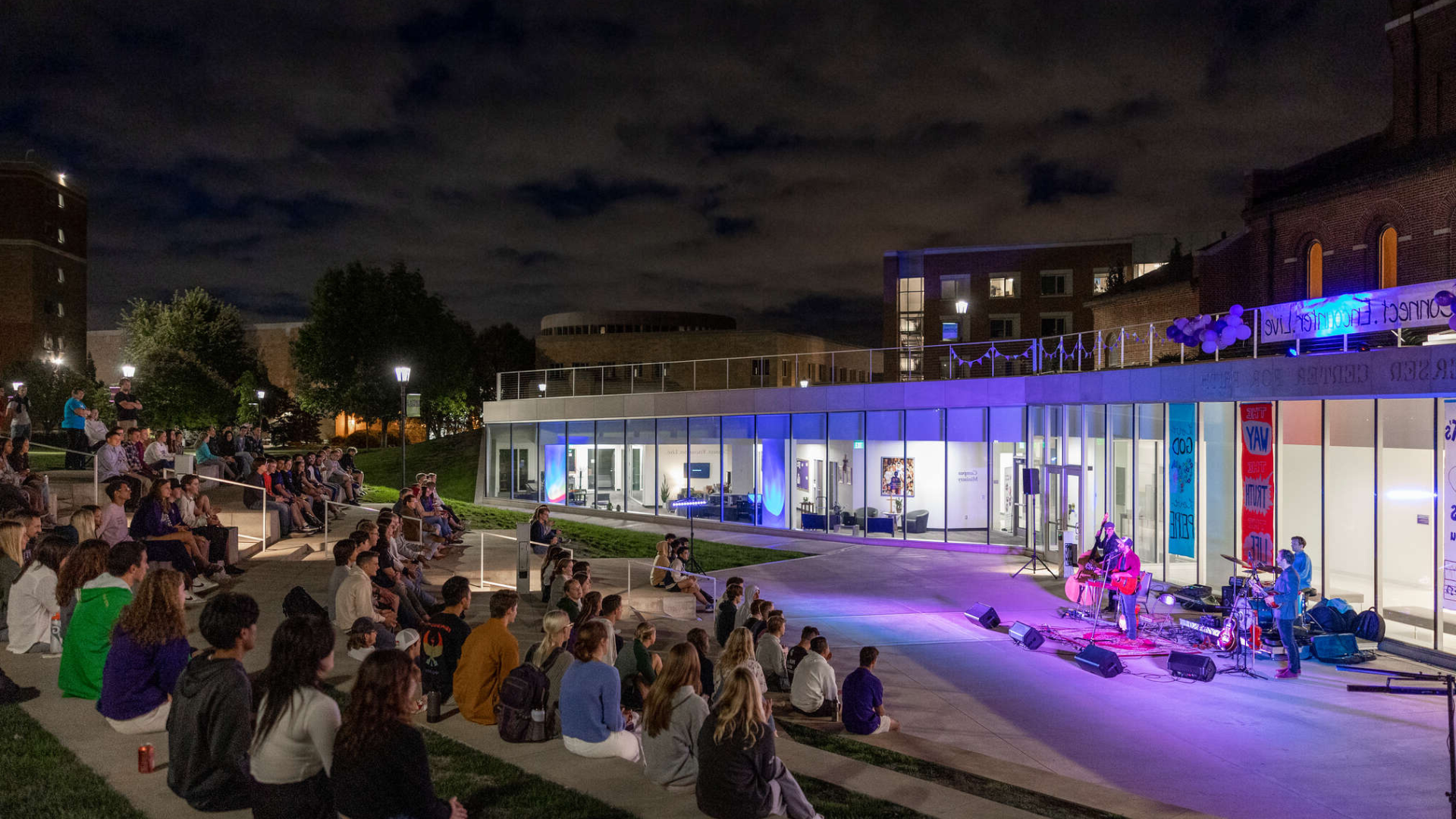 Students sit on amphitheater-style steps under the night sky, watching a live band perform outside a glass-front campus building lit in blue and purple lights. The event appears to be a campus gathering or block party with a relaxed and inviting atmosphere.