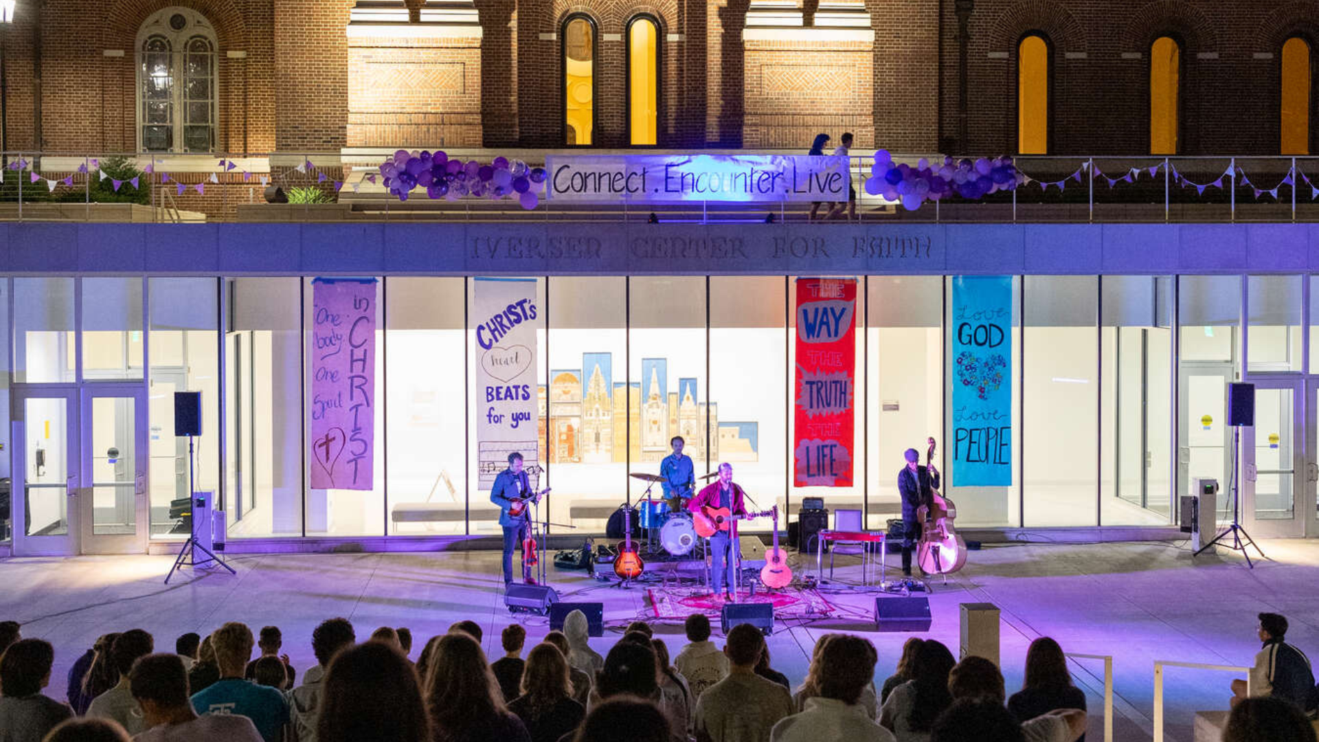 A band performs live outside the Iversen Center for Faith at night, illuminated by purple and blue lights. A crowd of students sits on the steps watching the musicians. Large banners in the background display spiritual messages such as “One body, One Spirit in Christ,” “Christ’s Beat Beats for You,” and “Love God, Love People.” Purple balloons and a banner reading “Connect. Encounter. Live” decorate the rooftop.