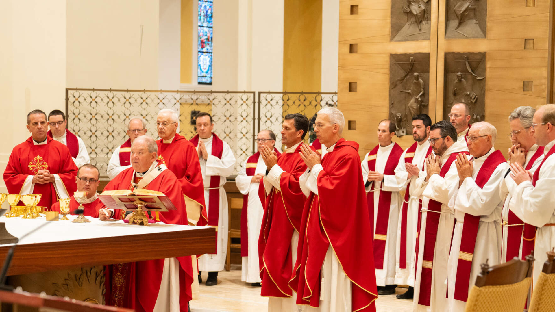 Catholic priests and deacons, dressed in red and white liturgical vestments, gather around the altar during the Mass of the Holy Spirit in a large, ornate chapel. A bishop at the altar leads the Eucharistic Prayer while others stand in reverent prayer behind him. Golden chalices and liturgical items are arranged on the altar.