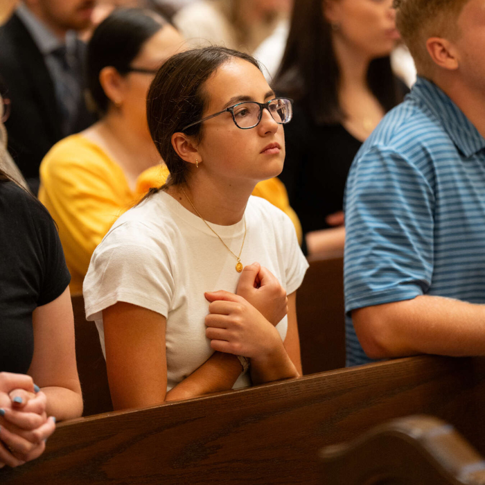 A young woman sits in a wooden pew with her hands crossed over her chest, appearing deep in prayer or reflection during a liturgical service. Other people are seated nearby in a warmly lit worship space.