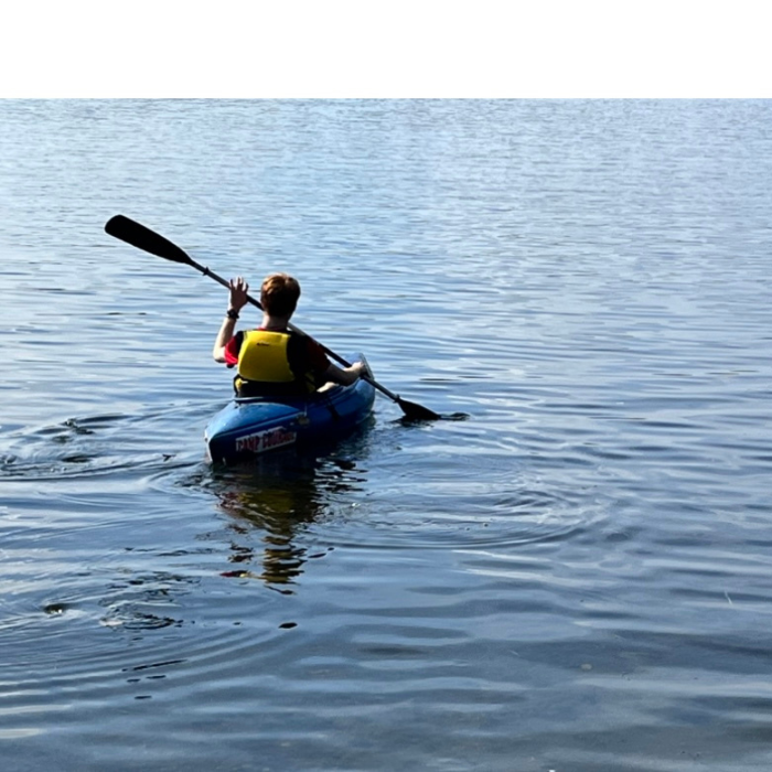 A person wearing a yellow life vest paddles a blue kayak alone on a calm lake, heading toward the open water under a clear sky. The peaceful scene suggests reflection and solitude, likely part of a retreat experience.