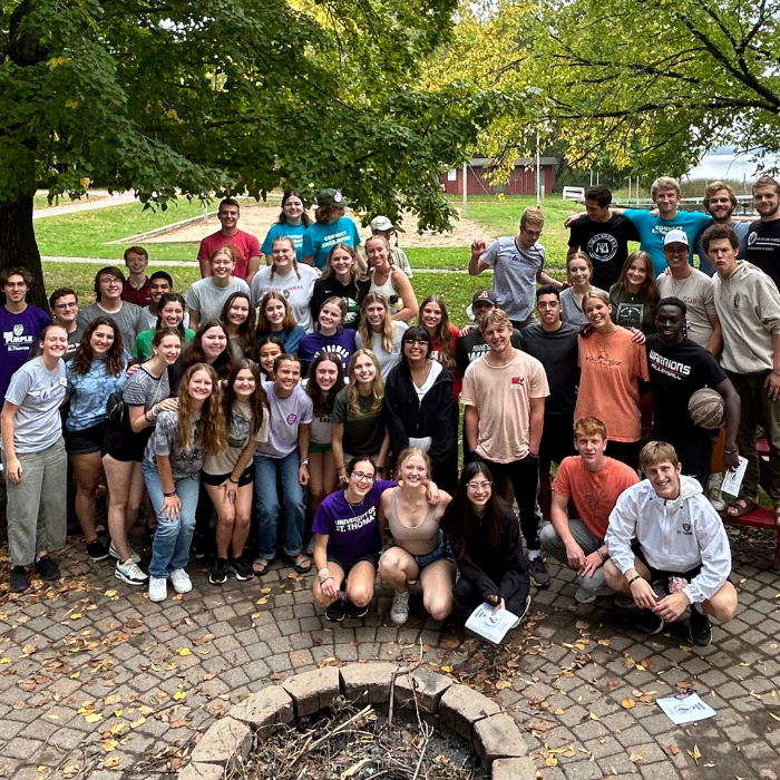 A large group of college students gathers for a joyful group photo outdoors during a retreat. They stand and kneel around a circular stone fire pit under leafy trees, smiling and dressed casually. The setting is peaceful with green grass, trees, and a red barn in the background.