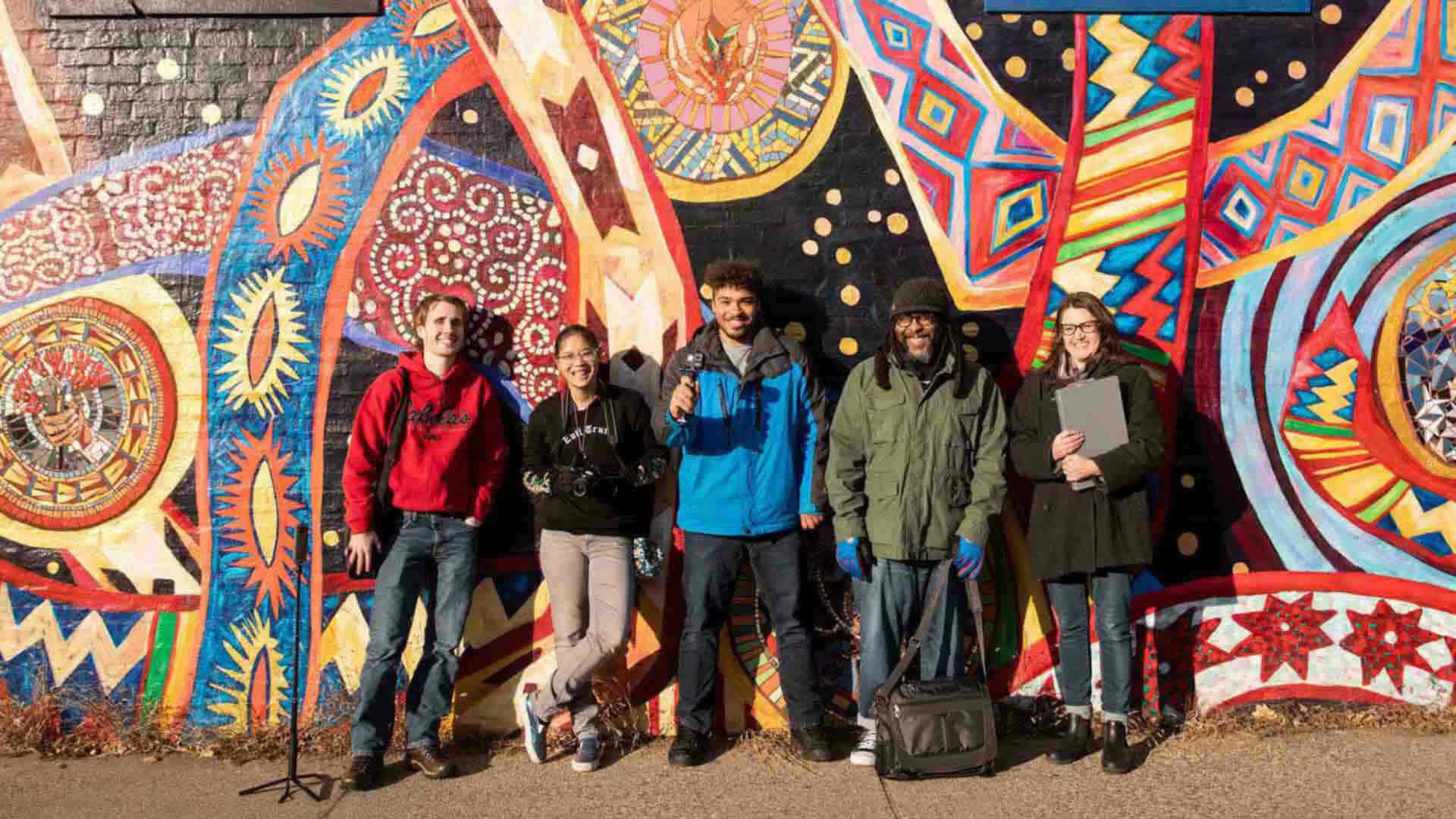 Student and faculty stand in front of a mural.
