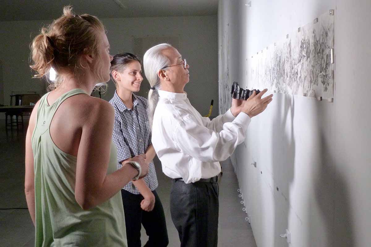 Three individuals analyze a piece of artwork displayed on a wall.