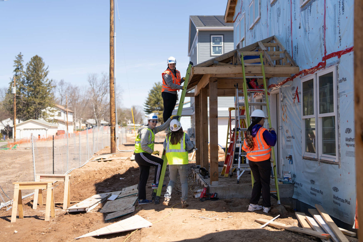 A group of student volunteers wearing hard hats and reflective vests work together to build a front porch on a Habitat for Humanity home. One student is on a ladder while others support and assist from the ground under a clear blue sky.