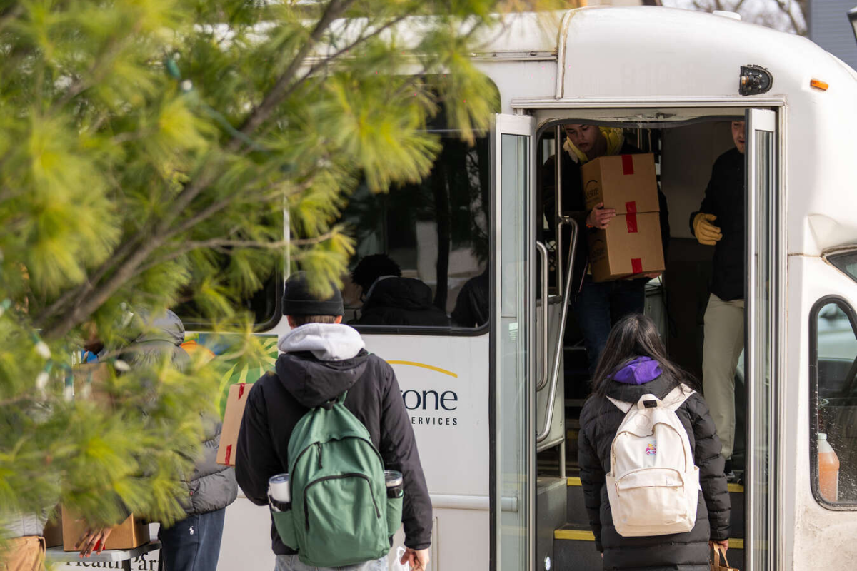 Volunteers unload cardboard boxes from a Keystone Community Services Foodmobile parked near campus. Students with backpacks wait in line, and team members inside the bus hand down boxes filled with food items.