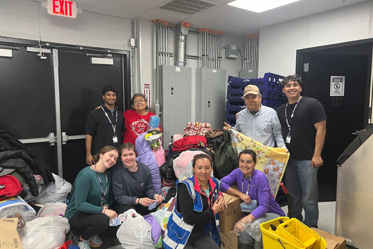 Students and community members pose together in a storage room filled with bags of donated clothing and blankets. They smile warmly after sorting and organizing supplies, demonstrating compassion and teamwork during a VISION trip.
