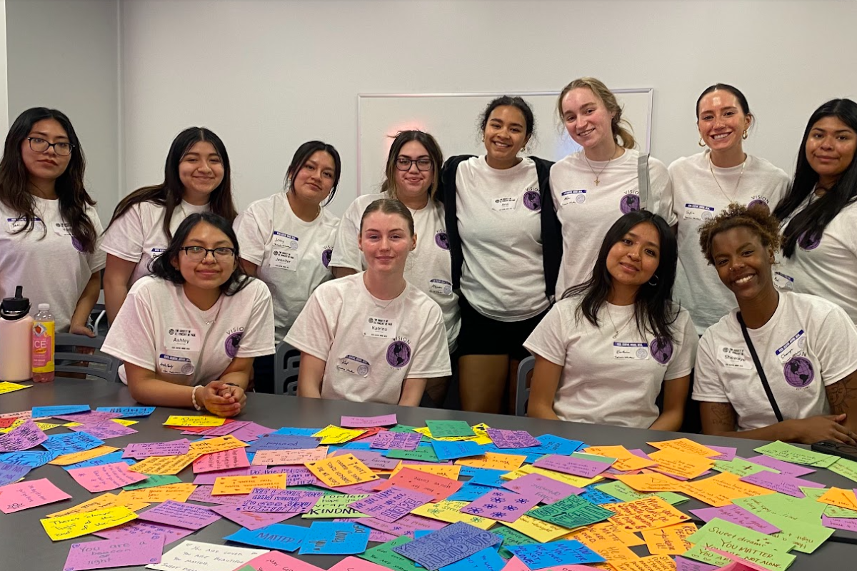 A group of VISION participants wearing matching white T-shirts pose around a table filled with colorful handwritten affirmation cards. The atmosphere is joyful and collaborative as they prepare messages of encouragement.