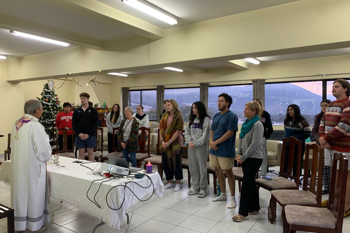 A small group of VISION students and community members stand in a semi-circle during a prayer service in a room decorated for Christmas. A priest leads the gathering near a table with a purple candle, a crucifix, and a projector, set against a windowed backdrop of mountains.