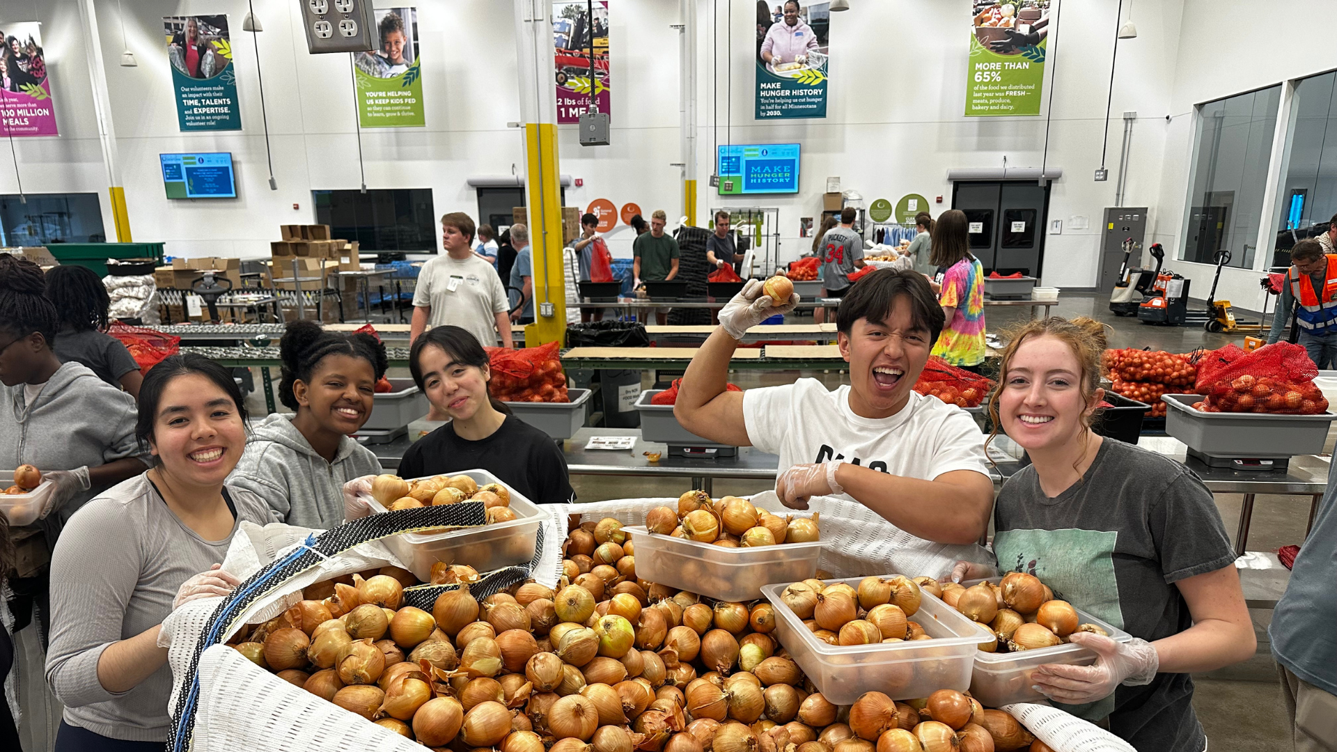 A group of six smiling young adults stands around a large bin full of onions, holding plastic containers and wearing gloves, inside a food distribution center. In the background, other volunteers are visible sorting produce under banners that promote hunger relief efforts.