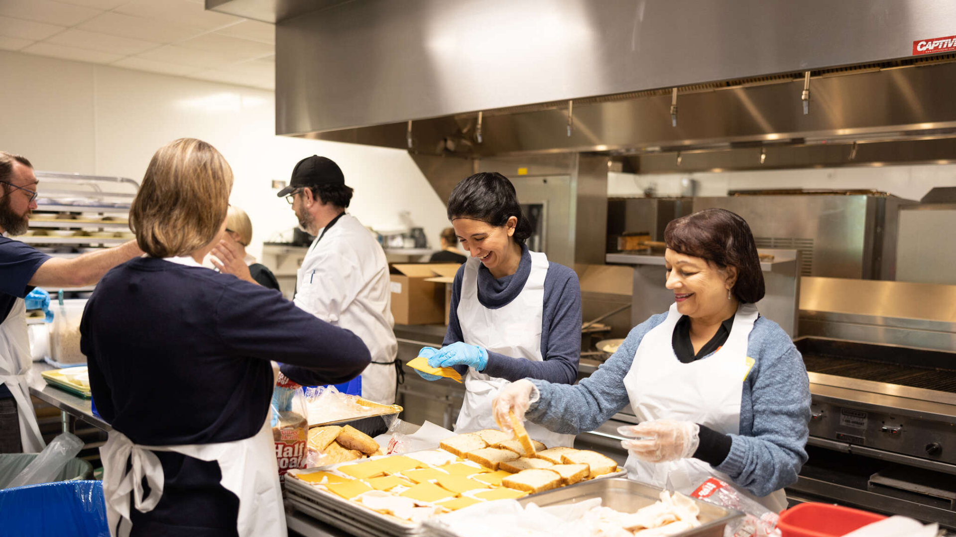Several students engage in painting a purple upright piano with colorful designs and words like “Evren Ozel” and “Eliana Elias.” The group is working in a sunlit room with paint supplies and orange buckets scattered on the floor.