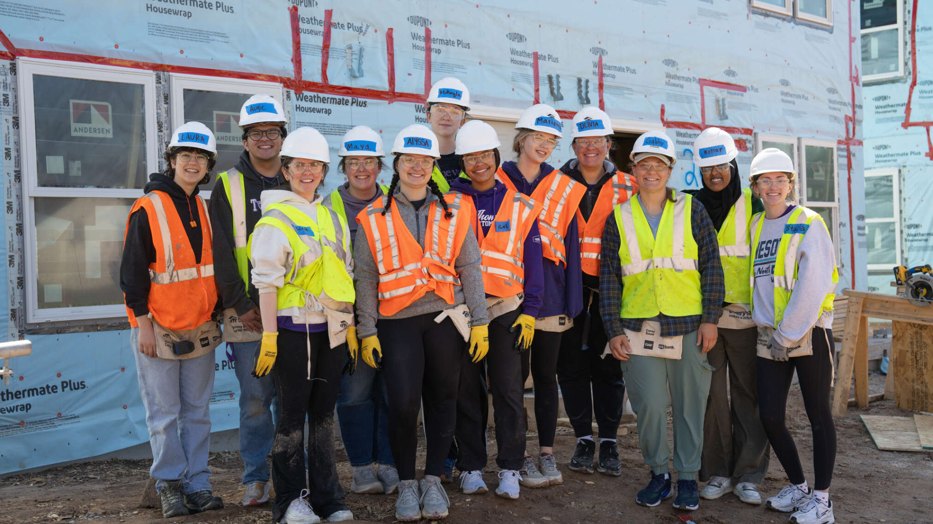 A group of students in hard hats, safety vests, and work gloves pose together at a construction site in front of a house under construction. They are participating in a Habitat for Humanity build, smiling and ready for service work.