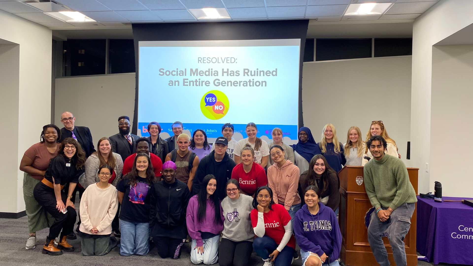 A group of students and facilitators smile together for a photo after a dialogue session. Behind them is a screen with the prompt: “Resolved: Social Media Has Ruined an Entire Generation” and YES/NO icons, indicating a structured conversation event.