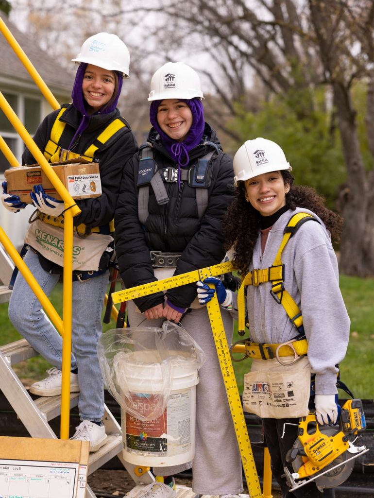 Three students wearing safety harnesses, hard hats, and Habitat for Humanity gear smile at the camera while standing near construction stairs. One holds a box of supplies, another a paint bucket and measuring stick, and the third a power saw, ready to work on a build site surrounded by trees in early spring.