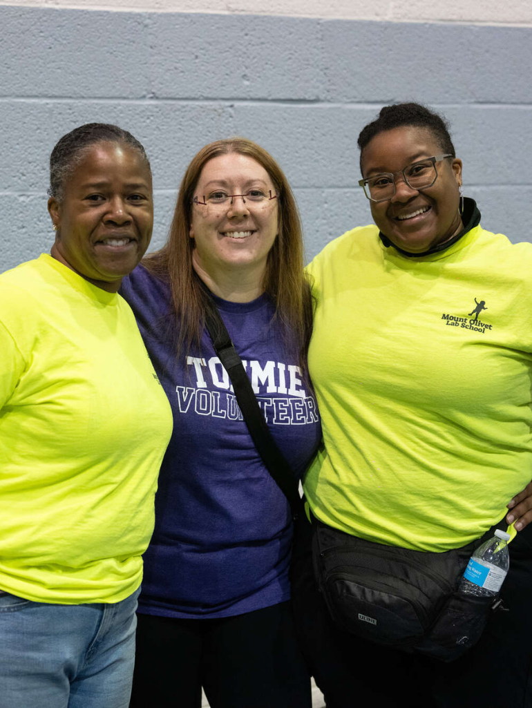 Three women smile together for a photo indoors, with one wearing a "Tommie Volunteer" t-shirt and the others in bright yellow "Mount Olivet Lab School" shirts. They stand closely, showing camaraderie during a volunteer event.