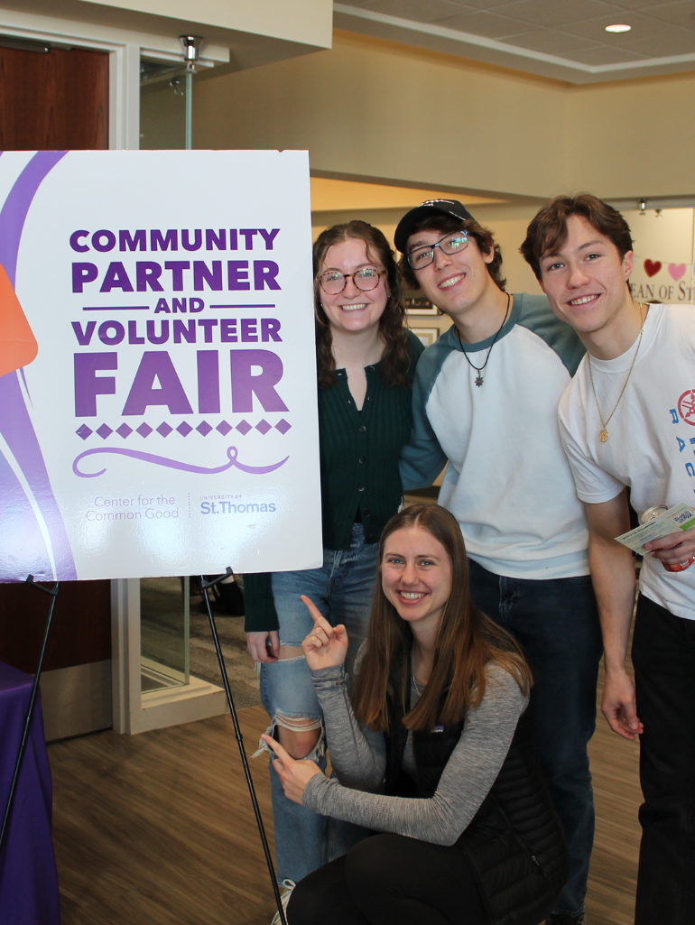 Four smiling students pose next to a large sign that reads “Community Partner and Volunteer Fair – Center for the Common Good – University of St. Thomas.” One student kneels and points at the sign, highlighting campus engagement with community partners.