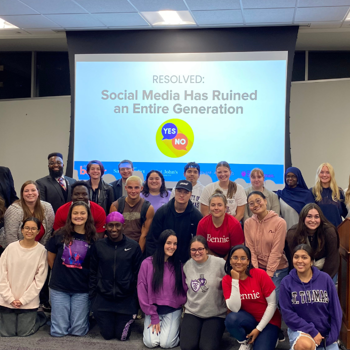 A group of students and facilitators smile together for a photo after a dialogue session. Behind them is a screen with the prompt: “Resolved: Social Media Has Ruined an Entire Generation” and YES/NO icons, indicating a structured conversation event.