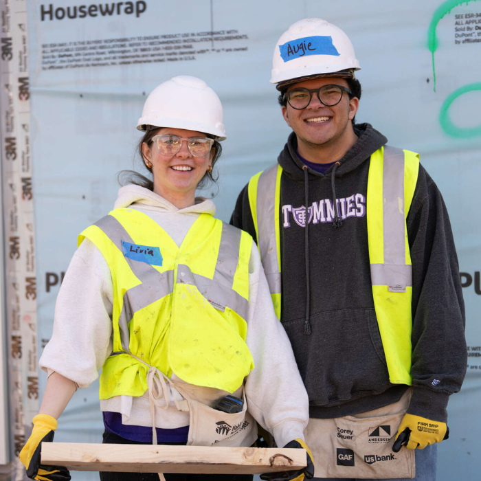 Two students in safety vests and hard hats stand smiling at a construction site. One holds a small plank of wood, and both are wearing Tommies apparel and protective eyewear while volunteering with Habitat for Humanity.