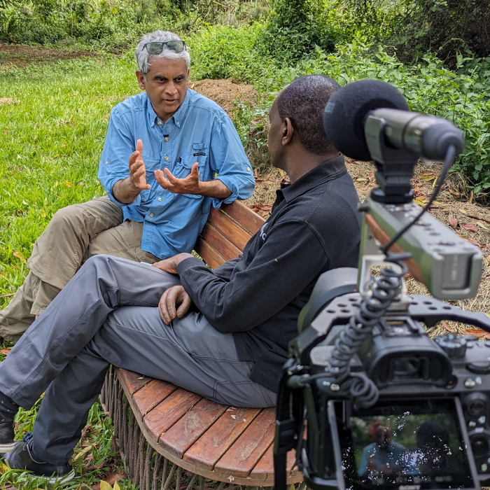 Two men sit on a curved wooden bench in a lush green outdoor setting having a filmed conversation. One gestures while speaking, and the other listens intently. A professional camera with a microphone is visible in the foreground recording the exchange.