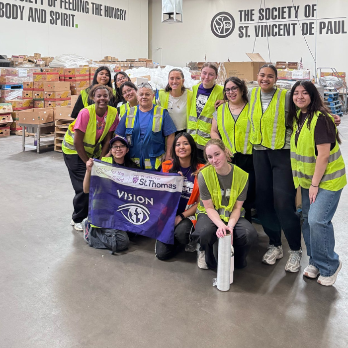 A group of student volunteers wearing reflective vests pose inside a warehouse at The Society of St. Vincent de Paul. They hold a purple VISION banner representing the University of St. Thomas and the Center for the Common Good.
