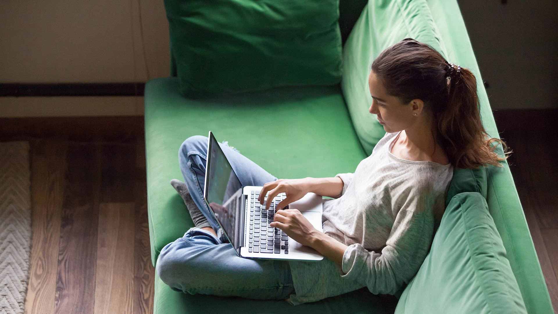 Student sits on a green couch and types away on her laptop.