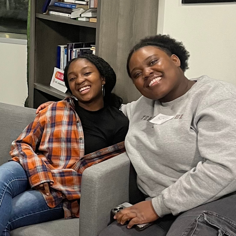 Two Black students sit close together on a couch, smiling brightly. One wears a gray sweatshirt and the other a plaid shirt layered over a black top. Bookshelves and a warm, inviting environment surround them.