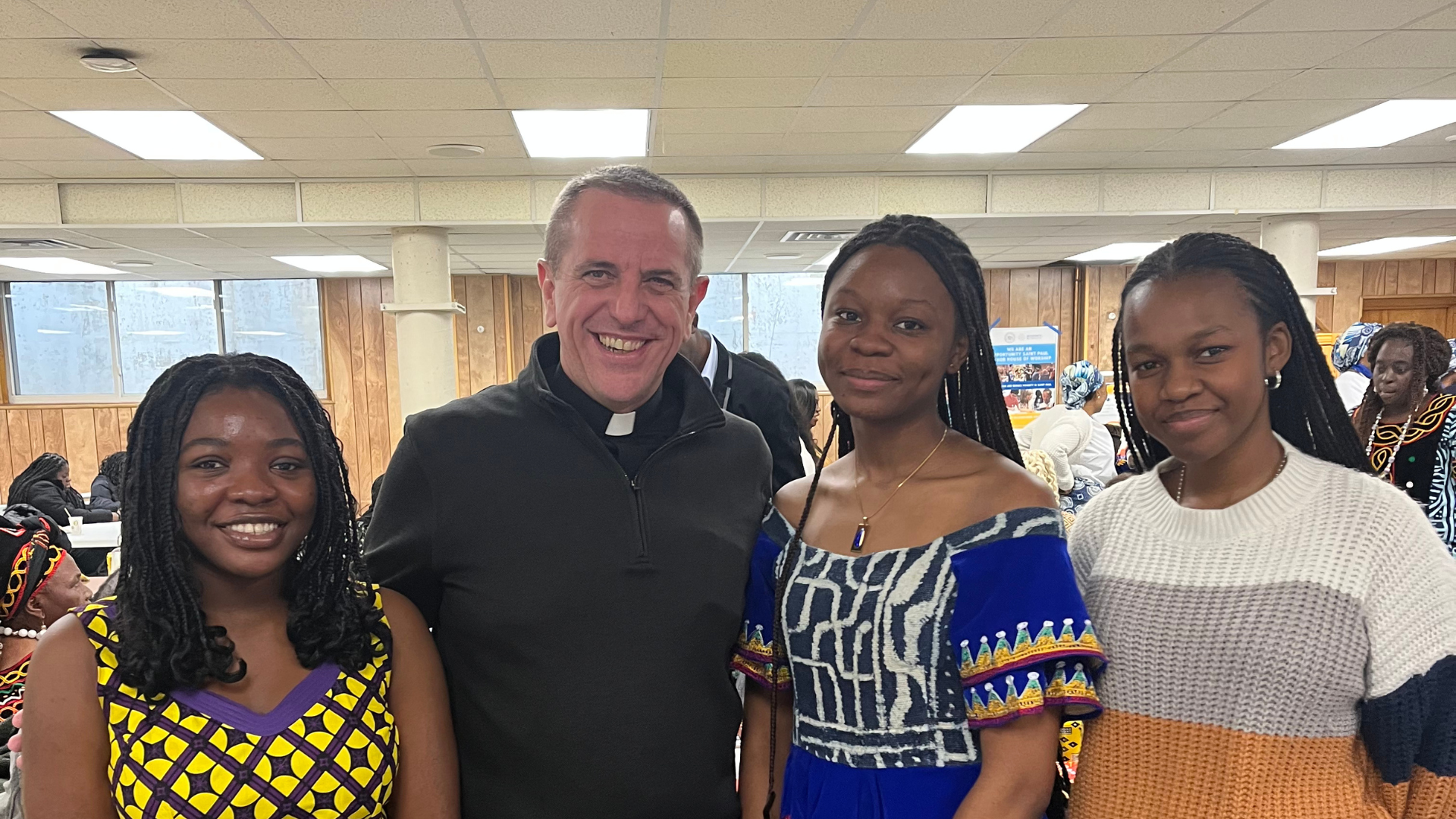 A Catholic priest smiles alongside three young women, two of whom wear colorful African dresses, while the third wears a cozy striped sweater. The group poses together during a joyful cultural or faith-centered event.