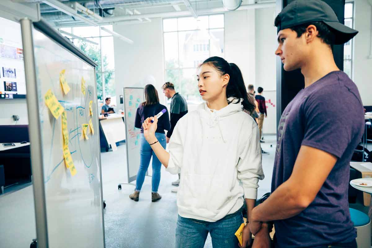 Two students work on exercises on a whiteboard in the Innovation Space in the Facilities and Design Center. 