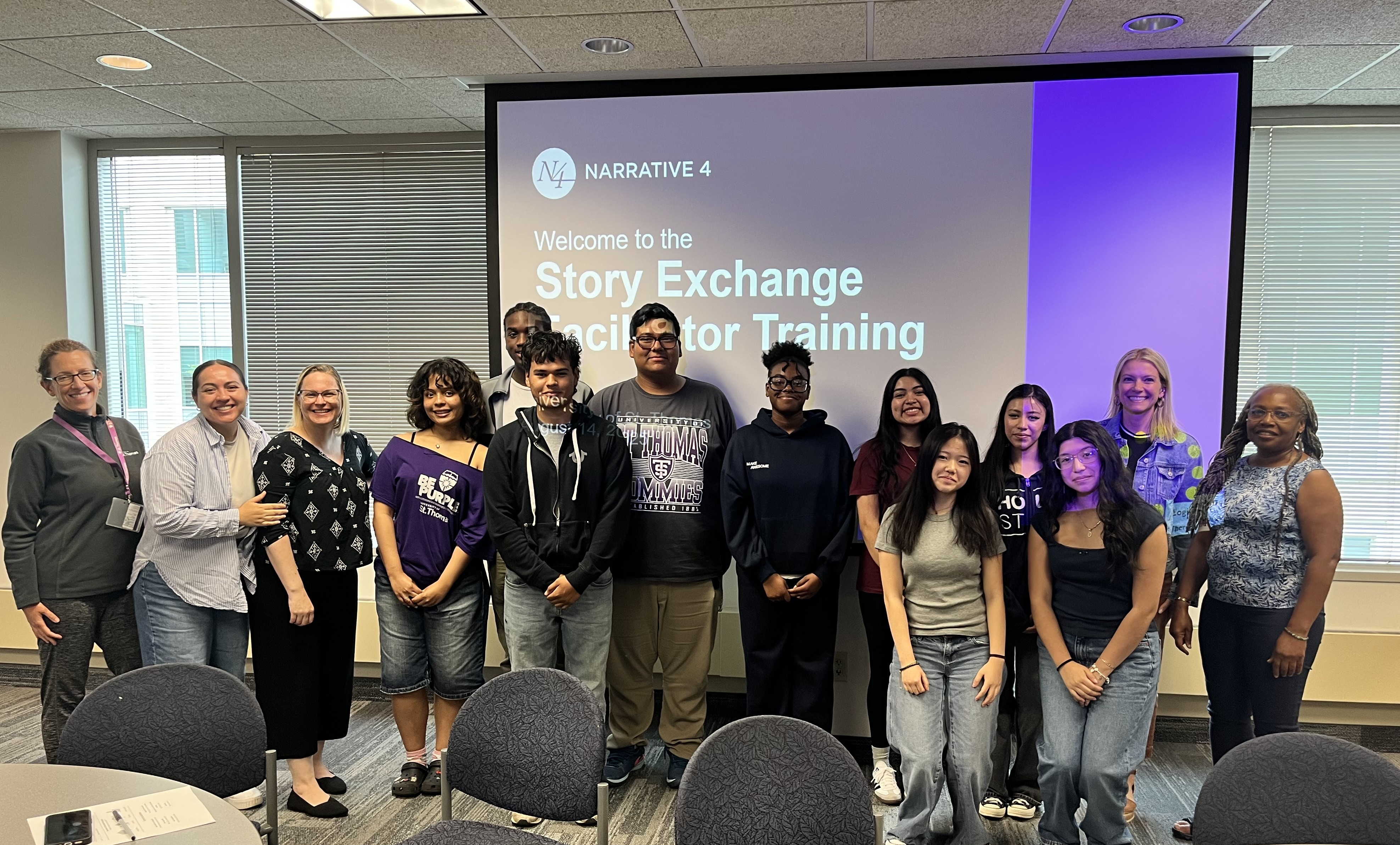 A group of students and facilitators stand together in a classroom, smiling for the camera. Behind them, a projector screen displays the title slide: “Narrative 4 – Welcome to the Story Exchange Facilitator Training.” The group includes about a dozen people of diverse ages and backgrounds, standing in two rows. The setting is a bright room with large windows and round tables with chairs in the foreground.